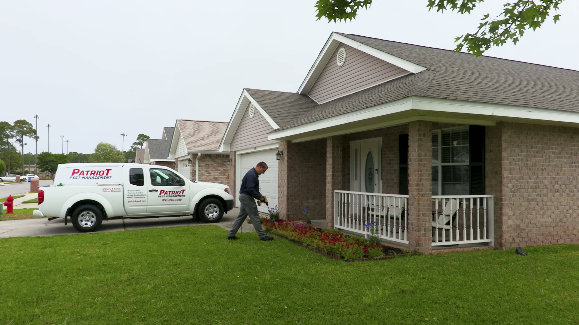 Pest control technician applying treatment to flower bed at brick home with white porch